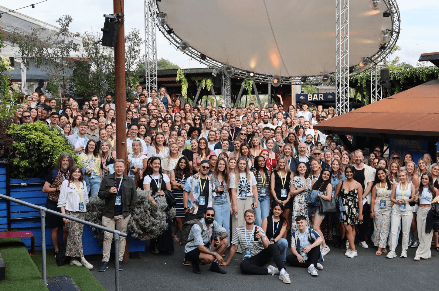 A large group photo of SMG employees gathered outdoors under a canopy at a company event. Everyone is smiling and looking at the camera, with some people holding drinks. The setting includes greenery, a bar sign in the background, and a festive atmosphere.