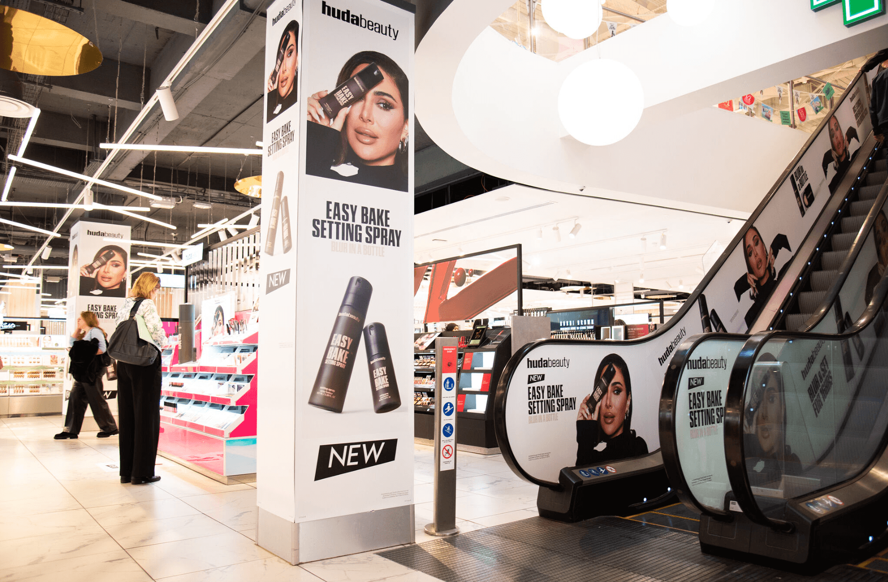 In-store beauty retail activation for Huda Beauty Easy Bake Setting Spray. Large posters, escalator wraps, and column signage display oversized product bottles and promotional images of a model with the slogan “Blur in a Bottle.” Shoppers are seen browsing nearby displays.